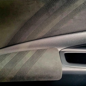 May include: Close-up of a car door interior. The door panel is a dark gray with a textured, suede-like material. Diagonal lines of small perforations are visible on the armrest and upper panel. The door handle and window controls are also visible.