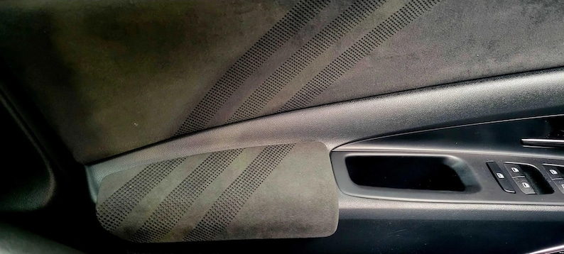 May include: Close-up of a car door interior. The door panel is a dark gray with a textured, suede-like material. Diagonal lines of small perforations are visible on the armrest and upper panel. The door handle and window controls are also visible.