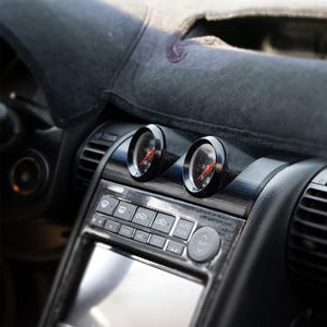 以下が含まれることがあります： 赤色の針が付いた2つの黒いゲージを備えた車のダッシュボードのクローズアップ。ダッシュボードは、さまざまなボタンと画面を備えた光沢のある黒い仕上げです。ゲージの上には濃い灰色のカバーがあります。