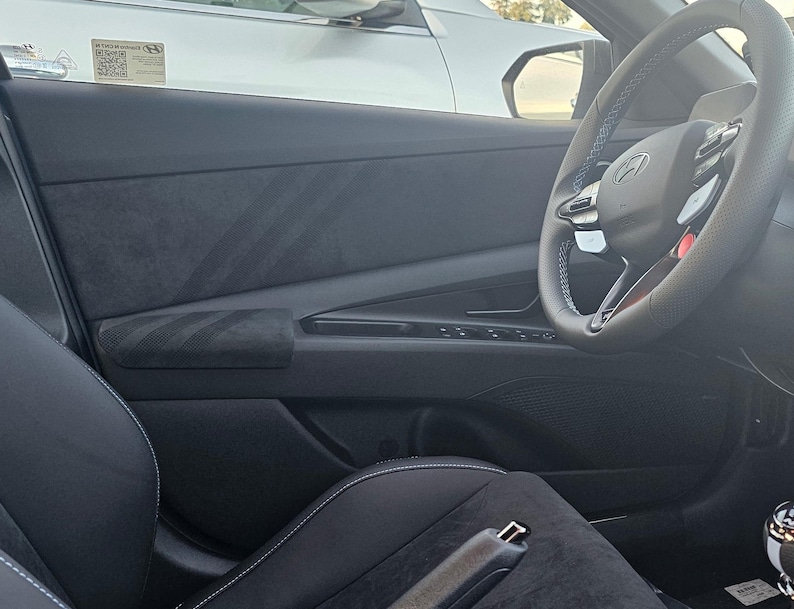 May include: Interior view of a car's front seat, featuring a black leather steering wheel with blue stitching and a red accent. The door panel is dark gray with a textured armrest. The seat is black leather with white stitching.