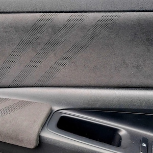 May include: Close-up of a car door panel featuring a dark gray suede-like material. The panel has a perforated design with diagonal stripes and a built-in armrest. The door handle and window controls are visible.