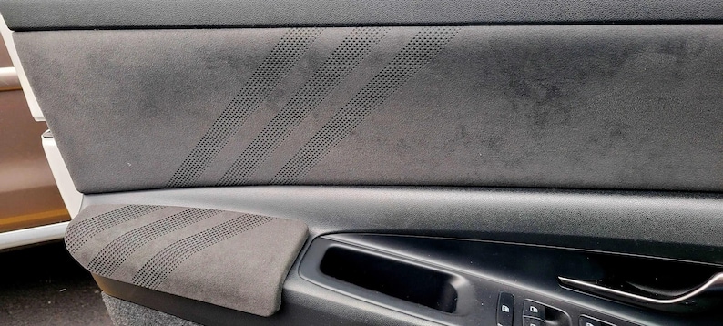 May include: Close-up of a car door panel featuring a dark gray suede-like material. The panel has a perforated design with diagonal stripes and a built-in armrest. The door handle and window controls are visible.