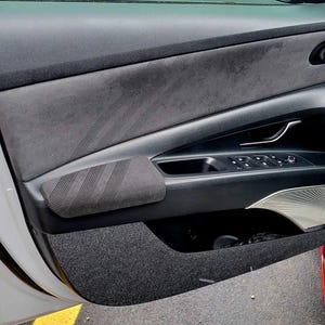May include: Interior car door panel with a gray suede-like upper section and a black lower section. The door features a black armrest with a striped design, control buttons, and a silver accent. The door is open, revealing a glimpse of the car's interior.