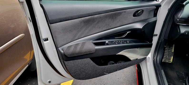 May include: Interior car door panel with a gray suede-like upper section and a black lower section. The door features a black armrest with a striped design, control buttons, and a silver accent. The door is open, revealing a glimpse of the car's interior.