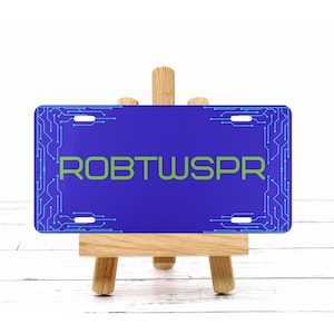 Op de afbeelding: Een blauwe nummerplaat met de tekst "ROBTWSPR" in het groen. De plaat heeft een printplaatontwerp langs de randen. De plaat wordt getoond op een klein houten schildersezel.