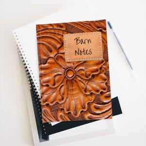 May include: A brown leather journal with a floral design and the words "Barn Notes" on a rectangular patch. The journal is on top of a stack of notebooks and a pen.