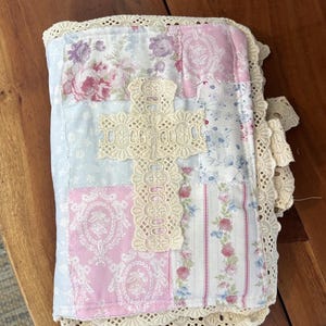 May include: A rectangular fabric cover featuring a patchwork design of pink, blue, and white floral and striped patterns. A cream-coloured lace cross is centred on the front, and the entire perimeter is finished with a wide, scalloped lace trim.