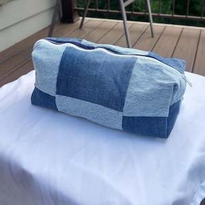 Op de afbeelding: Een rechthoekige tas met ritssluiting, gemaakt van een patchwork van denim stoffen vierkanten in afwisselende tinten licht- en middenblauw. De tas heeft een witte ritssluiting aan de bovenkant en een gestructureerde, hoekige vorm.