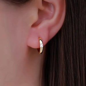 May include: A close-up of a gold hoop earring with a small, square, clear gemstone. The earring is worn on a person's ear. The earring is a simple, elegant design.
