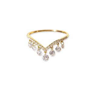 May include: A gold-colored ring with a V-shaped design. The ring is adorned with small, sparkling clear stones along the top and dangling clear stones. The ring is set against a white background.