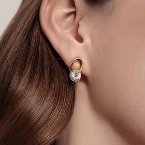 May include: Gold-tone earrings with a clear, round, sparkling gemstone. The earring design features a gold loop that encircles the gemstone. The earrings are shown on a person's ear, highlighting the jewelry's design and sparkle. A simple and elegant accessory.