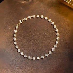 Può includere: Un bracciale a catena dorata caratterizzato da una serie di pietre rotonde, trasparenti e sfaccettate, incastonate in castoni dorati individuali. Il bracciale è collegato da piccoli anelli dorati e include una chiusura a moschettone.