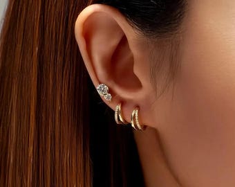 May include: A close-up of an ear adorned with multiple gold earrings. The ear has two small stud earrings with clear stones and two sets of small gold hoop earrings. The earrings are set against the backdrop of long, brown hair.