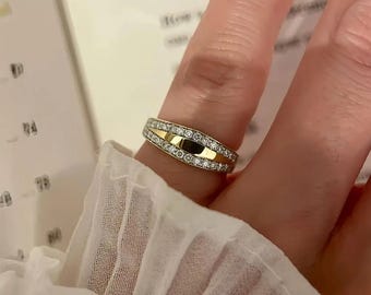 Może przedstawiać: Złoty pierścionek z diamentami. Pierścionek ma centralny złoty pas otoczony rzędami małych, błyszczących diamentów. Design jest prosty, a zarazem elegancki, odpowiedni do noszenia na co dzień lub na specjalne okazje.
