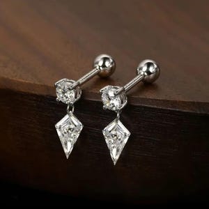 May include: A pair of silver earrings. Each earring has a round stud and a clear, diamond-shaped pendant. The earrings are presented on a dark wooden surface.