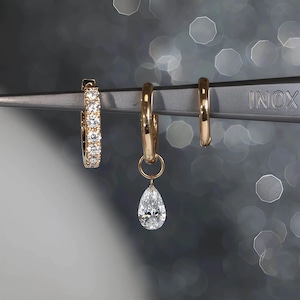 May include: Three gold earrings displayed on a metal tool. One earring is a hoop with embedded clear stones, another is a simple gold hoop, and the third features a teardrop-shaped clear stone pendant. The background is a blurred, bokeh effect.