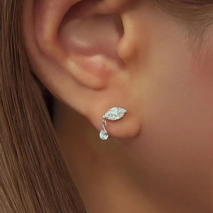 May include: A close-up of a diamond earring on an ear. The earring features a marquise-cut diamond and a dangling pear-shaped diamond. The earring is set in a gold metal.