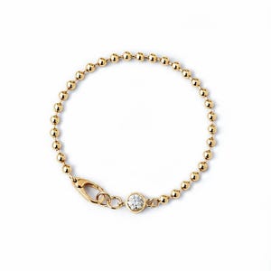 Könnte beinhalten: Ein goldfarbenes Kugelkettchen-Armband mit einem einzelnen runden, klaren, facettierten Stein in einer Zargenfassung. Das Armband verfügt über einen Karabinerverschluss, der mit der Steinfassung verbunden ist und ein kreisförmiges Design bildet.