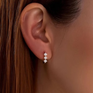 Könnte beinhalten: Nahaufnahme eines goldenen Ohrrings mit drei kleinen, klaren Edelsteinen. Der Ohrring ist an einem Ohr befestigt, wobei die Edelsteine vertikal angeordnet sind.