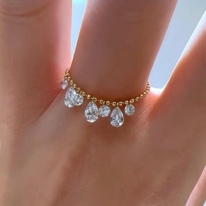 May include: A gold ring with a series of dangling, teardrop-shaped and round clear gemstones. The ring is worn on a finger, highlighting the intricate design and sparkle of the gemstones. The ring's band is decorated with small, round gold beads.