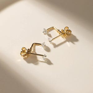 Könnte beinhalten: Ein Paar goldfarbene Ohrringe mit L-förmigem Stecker. Jeder Ohrring hat einen kleinen, klaren, facettierten Stein am Ende des Steckers und einen weiteren klaren Stein an der Ecke des L-Stücks, befestigt mit einem goldfarbenen Schmetterlingsverschluss.