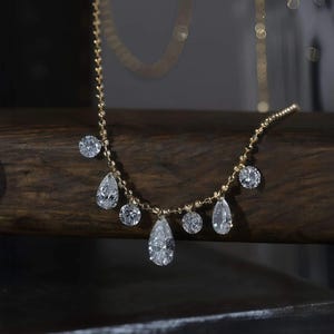 Può includere: Una delicata collana d'oro con pendenti di diamanti trasparenti, di forma rotonda e a goccia. La collana è esposta su una superficie di legno scuro, che mette in risalto la brillantezza delle pietre preziose.