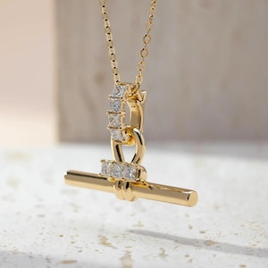 Può includere: Una collana color oro con un ciondolo a forma di chiusura a T. Il ciondolo è composto da una barra orizzontale collegata a un anello circolare, entrambi decorati con piccole pietre trasparenti a taglio quadrato, su una sottile catena dorata.