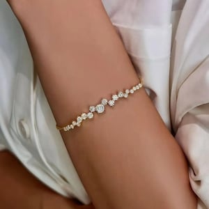 May include: A delicate gold bracelet featuring a cluster of sparkling, round-cut gemstones. The bracelet is designed with a series of varying sized stones, creating a unique and elegant look. The bracelet is shown on a person's arm, against a white shirt.