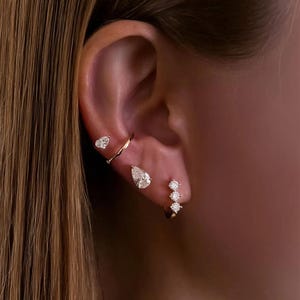 Puede incluir: Primer plano de una oreja adornada con múltiples pendientes dorados. Se ve un pendiente en forma de corazón, un delicado ear cuff, un pendiente en forma de lágrima y un pequeño aro con tres piedras brillantes.