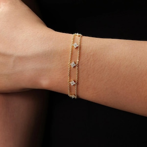 Pode incluir: Uma pulseira de corrente dupla em tom dourado com quatro berloques em forma de flor espaçados uniformemente. Cada berloque é incrustado com pequenas pedras claras e brilhantes. As delicadas correntes de metal conectam os motivos florais.