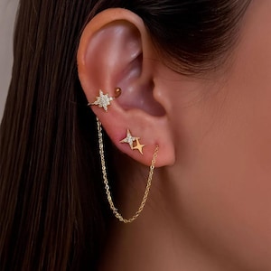May include: Gold-toned ear jewelry set featuring a star-shaped ear cuff with clear crystals, a stud earring, and a chain connecting the two. The jewelry is worn on the ear, showcasing the design and details.