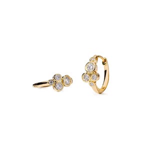 May include: A pair of gold hoop earrings, each featuring a cluster of four round, clear, bezel-set stones arranged in a tiered formation along the front of the hoop. The metal has a polished, reflective finish, and the stones vary slightly in size.