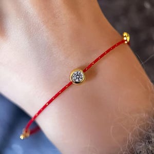 May include: A red string bracelet featuring a gold bezel-set diamond. The bracelet has gold detailing at the ends. The diamond is round and clear, set in a gold bezel.