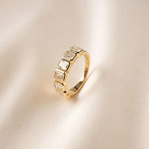Könnte beinhalten: Ein Goldring mit fünf klaren Steinen im Smaragdschliff, die in einer Reihe angeordnet sind. Die Steine werden von einzelnen Goldfassungen gehalten, die ein durchgehendes Band bilden. Das Metall hat eine polierte, reflektierende Oberfläche.