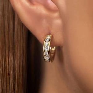 Può includere: Un orecchino a cerchio dorato con una fila di pietre chiare e scintillanti. L'orecchino è indossato sull'orecchio, mettendo in risalto il design e il modo in cui cattura la luce. Questo gioiello è un accessorio classico ed elegante.