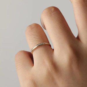 Könnte beinhalten: Ein dünner Goldring mit einer Kanalfassung, bei der eine durchgehende Reihe kleiner, klarer, funkelnder Steine in die Außenfläche eingelassen ist. Das Metall hat eine polierte, reflektierende Oberfläche, die das Licht einfängt.