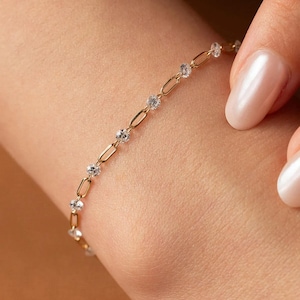May include: A gold chain bracelet featuring a series of alternating elongated oval links and small, clear, round-cut stones. The bracelet is worn around the wrist, showcasing a delicate design with a polished metallic finish and sparkling accents.