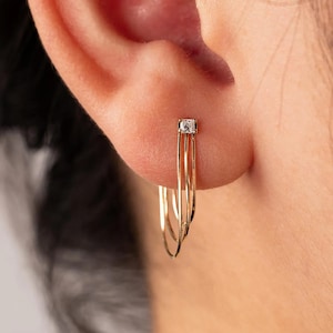 May include: A gold hoop earring featuring three thin, layered bands that converge at the top. A single, small, square-cut clear gemstone is set at the point where the three bands meet, securing the earring to the earlobe with a post mechanism.