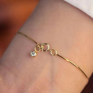 Könnte beinhalten: Ein zartes goldenes Armband mit einem kleinen, funkelnden Diamantanhänger. Das Armband hat eine dünne Kette und einen einfachen Verschluss. Der Diamant ist rund und in einer klassischen Lünettenfassung gefasst. Das Armband wird am Handgelenk gezeigt.