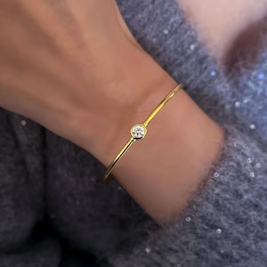 May include: A gold-colored bangle bracelet with a single, round, clear gemstone. The bracelet is thin and delicate, with the gemstone set in a simple bezel. The bracelet is worn on a wrist, against a gray sweater.