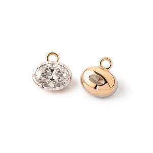 Puede incluir: Dos dijes ovalados para joyería con una piedra central transparente facetada engastada en un bisel de metal dorado pulido. Cada dije incluye una pequeña anilla circular en la parte superior para su sujeción. La superficie es lisa.