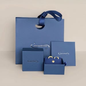 May include: A collection of jewelry packaging in shades of blue. The set includes a shopping bag with a ribbon handle, two boxes, and a small open box displaying gold hoop earrings. The brand name "Opreeshyne" is visible.