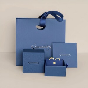 May include: A collection of jewelry packaging in shades of blue. The set includes a shopping bag with a ribbon handle, two boxes, and a small open box displaying gold hoop earrings. The brand name "Opreshyne" is visible.