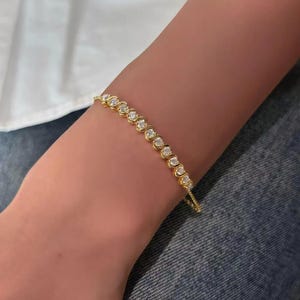May include: A gold bracelet with a series of round, clear gemstones. The bracelet is worn on a wrist, with a white shirt and blue jeans in the background. The bracelet is a delicate piece of jewelry.