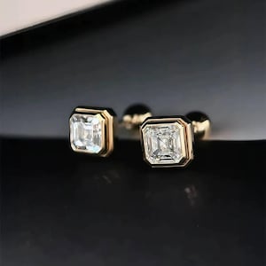 Op de afbeelding: Een paar vierkant geslepen diamantstud-oorbellen met een gouden bezel-zetting. De diamanten zijn helder en sprankelend, gezet in een gouden frame. De oorbellen worden gepresenteerd op een zwart oppervlak.