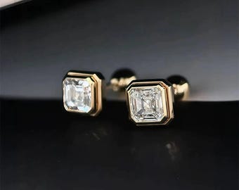 May include: A pair of square-cut diamond stud earrings with a gold bezel setting. The diamonds are clear and sparkling, set in a gold frame. The earrings are displayed on a black surface.