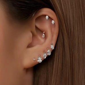 Puede incluir: Primer plano de una oreja adornada con múltiples pendientes de oro y diamantes. Los pendientes incluyen pequeños tachuelas, diseños en forma de lágrima y marquesa, creando un aspecto elegante y en capas. La oreja se encuentra sobre un fondo de cabello castaño.