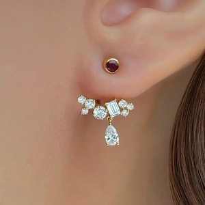 May include: Close-up of a gold earring set with multiple clear, sparkling gemstones. The design includes a cluster of round stones, a rectangular stone, and a dangling pear-shaped stone. A small, round stud earring with a red stone is also visible.