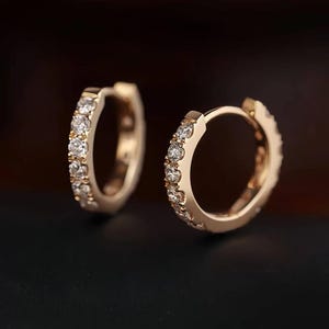 Puede incluir: Un par de pendientes de aro dorados, cada uno incrustado con pequeñas piedras preciosas transparentes. Los pendientes se muestran sobre un fondo oscuro, resaltando sus detalles brillantes. El diseño es simple pero elegante, adecuado para el uso diario o ocasiones especiales.