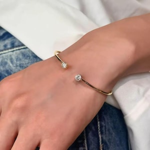 May include: A gold-colored cuff bracelet with a sparkling, round, clear gemstone at each end. The bracelet is worn on a hand, against a backdrop of a white shirt and blue denim.
